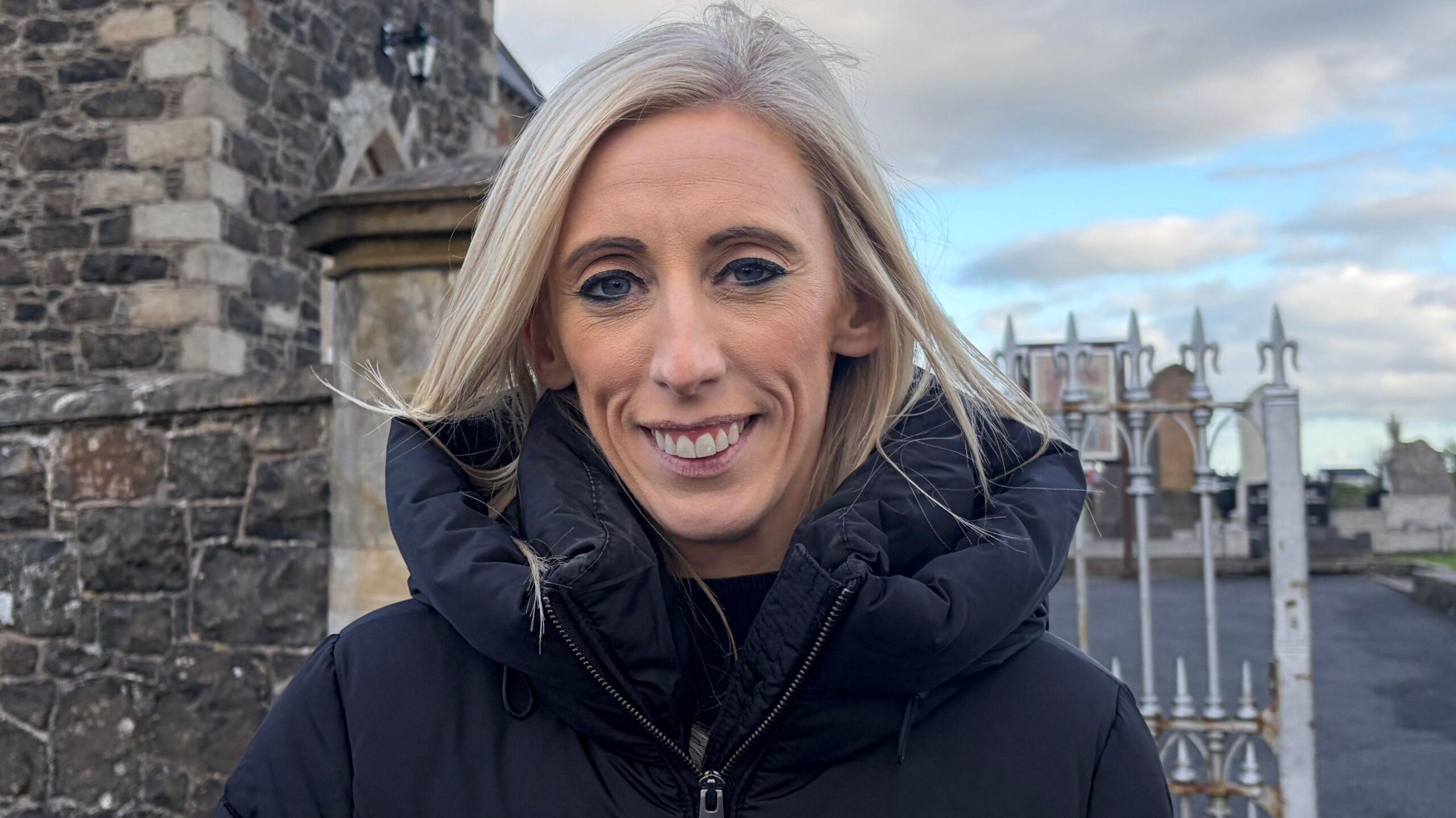 A woman with blonde hair smiles at the camera, she has a black coat on
