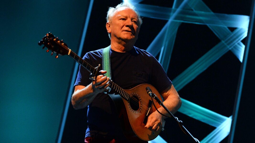 A photo of a man with grey hair, in a navy tshirt, playing a guitar on stage