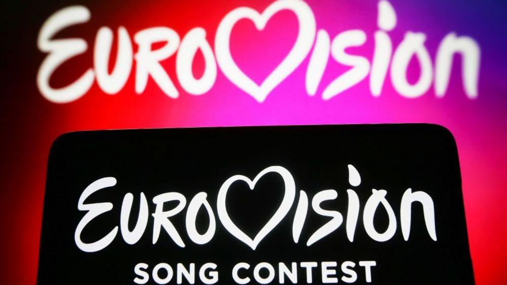 Two Eurovision backdrops, one with white words on a pink and red background and the other with white words on a black background in the foreground.
