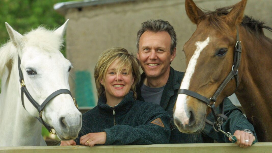 Sarah Fisher and Anthony Head standing in between two horses. The horse on the left is white and the horse on the right is brown with a white stripe down its nose. Sarah and Anthony are stood close together, looking at the camera and smiling.