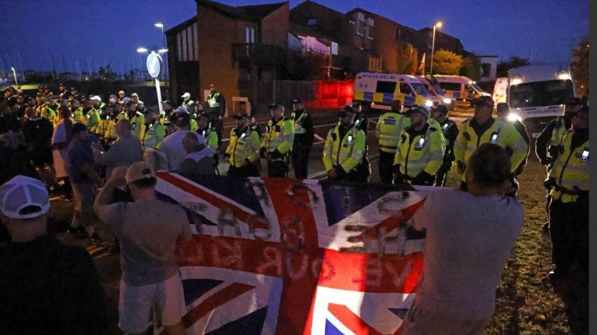 Police and demonstrators during a protest in Gosport.  A line of police officers are facing towards a crowd holding flags in a street - it is dark.