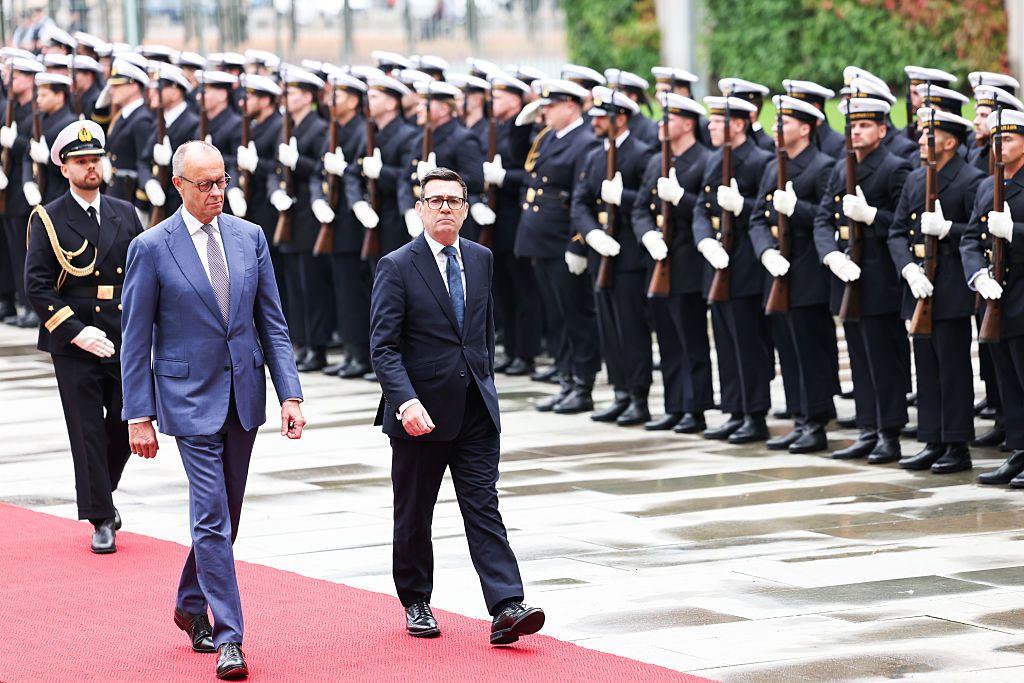 Merz and Burnham walk along a red carpet with rows of military personnel in the background, wearing ceremonial dress.