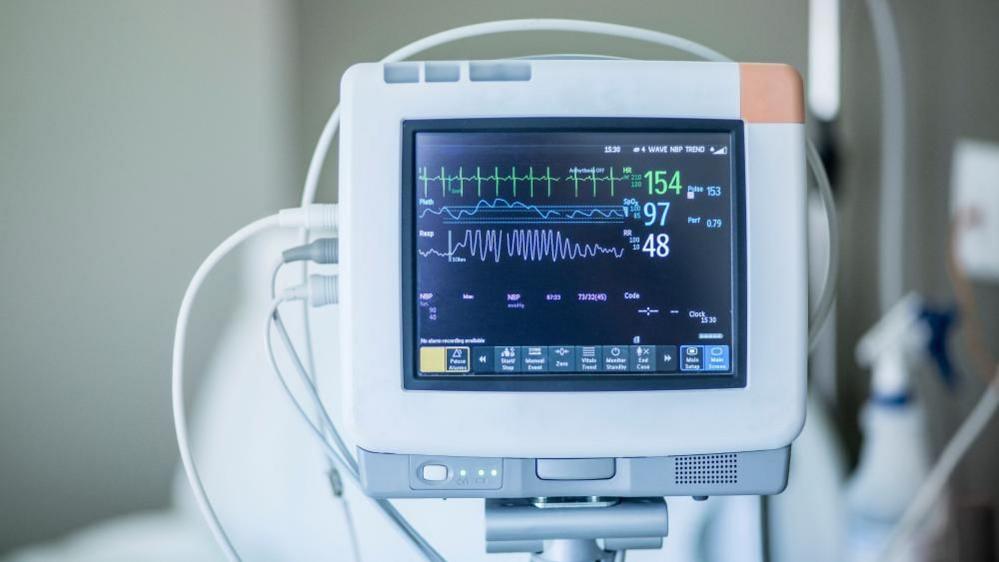 A monitor displays numbers and graphs for the key vital signs of a hospital patient. Three wires are attached to the device, which comprises a small, black screen inside a grey and white case. A blurred bed and bottle of cleaning spray feature in the background.