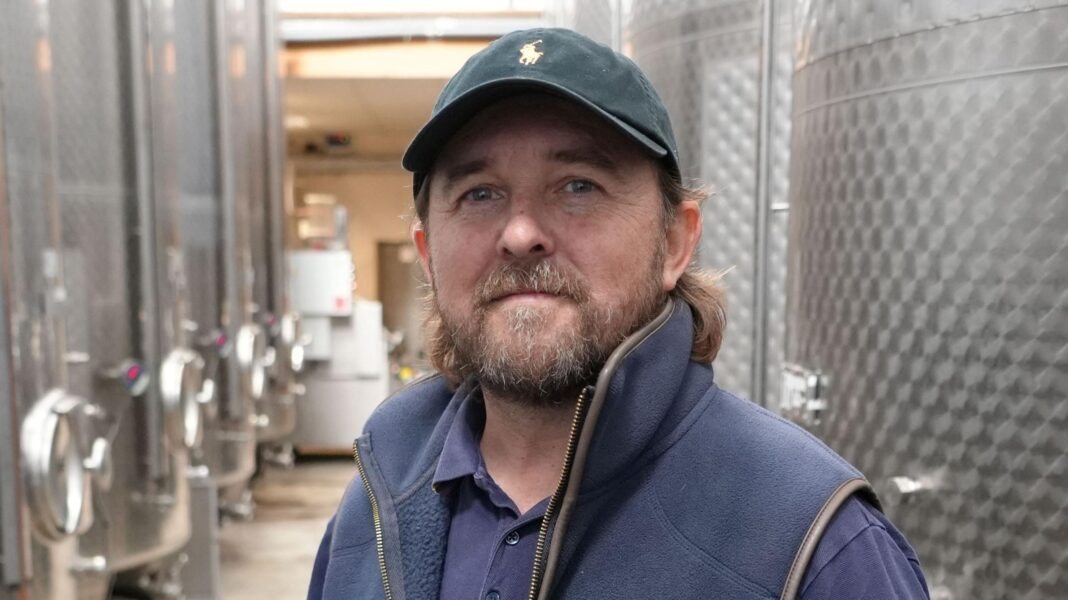 Lee Dyer standing inside in front of large metal vats. He is wearing a blue baseball cap, a blue gilet over a blue shirt. He has a beard and shoulder-length mid-brown hair.