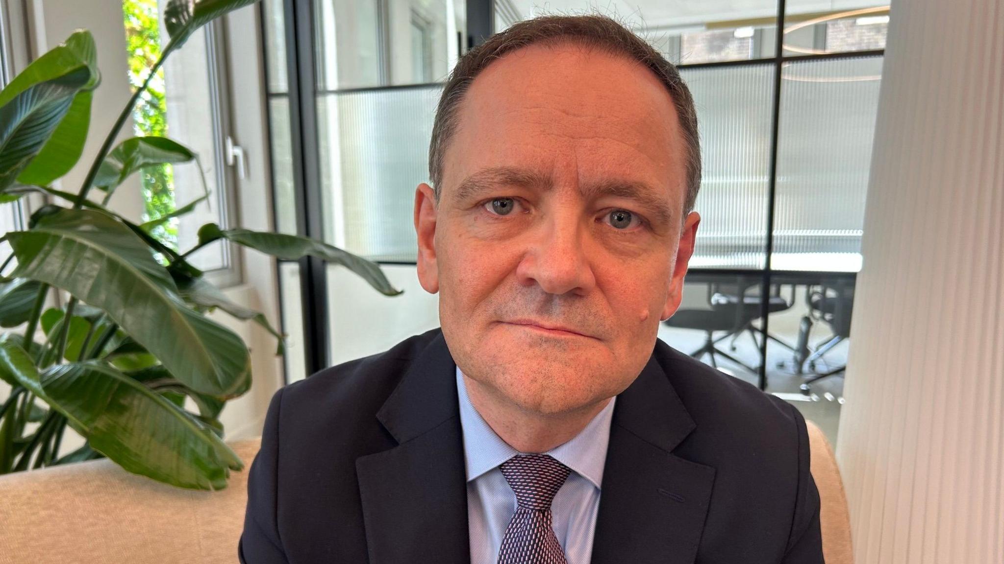 A man wearing a suit sat on a chair next to a plant looking intensely into the camera