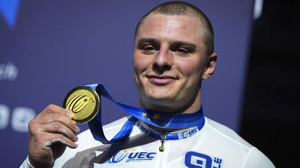 GB cyclist Matt Richardson holds up a gold medal while smiling at the camera