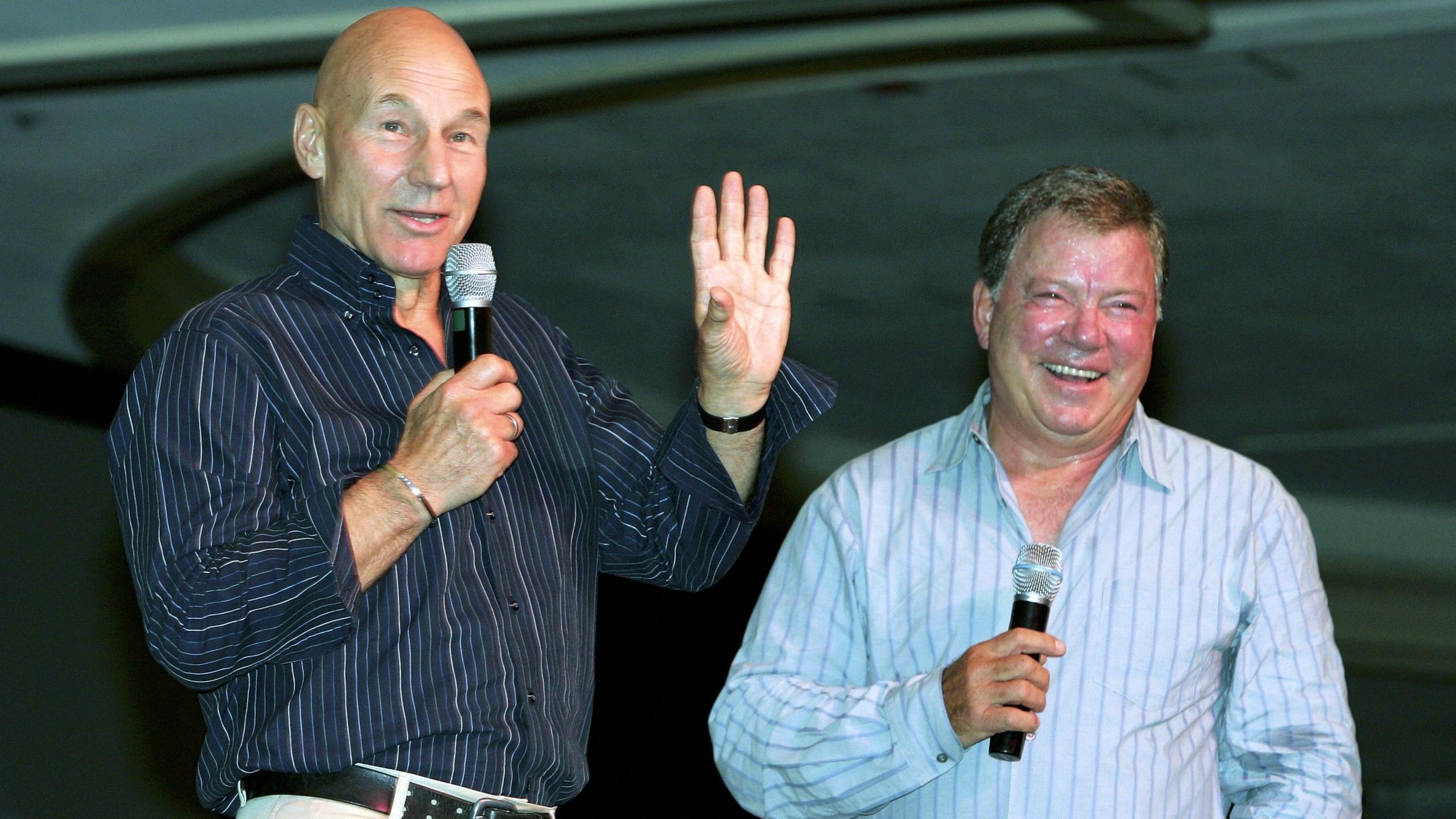Sir Patrick Stewart and William Shatner appear at a speaking event together in 2004. Both hold microphones and are wearing striped shirts.