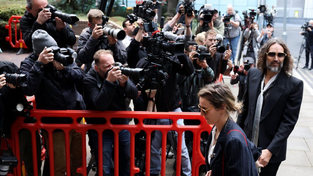 Russell and Laura Brand holding hands as they walk past a group of photographers behind a red barrier outside court