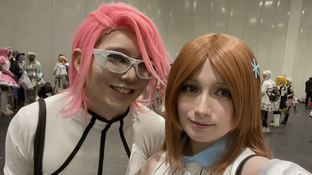 Nicholas on the right wears a white bodysuit with black piping and a pink wig. Ruby wears a blue collar and white striped outfit and a brown wig. They both look at the camera and smile.