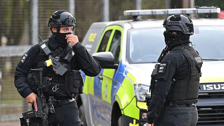Two armed counter-terror police maintain road blocks and security checks around RAF Fairford both are wearing full black uniforms, helmets and are carrying guns.