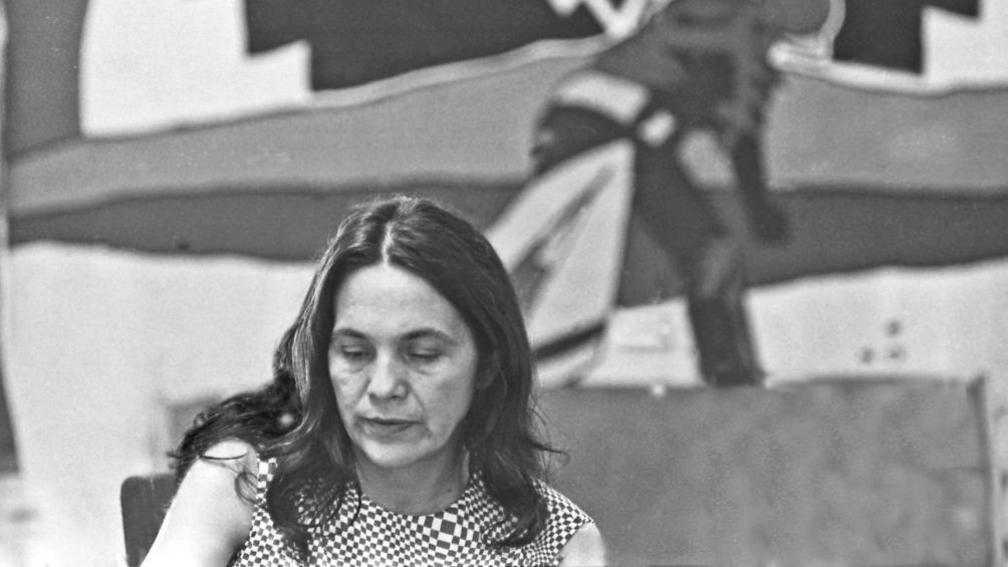 Black and white photo of Huerta with longer hair looking down whil wearing a checked top, in front of a mural of a person bending over, presumably to pick crops