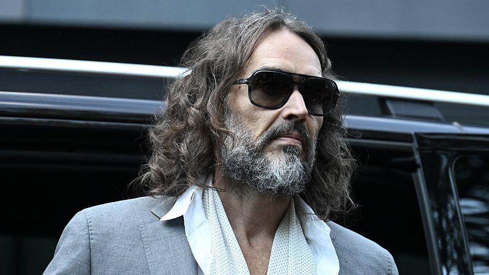 Russell Brand in a grey suit, white shirt and long, white scarf, leaving a car to enter the court room