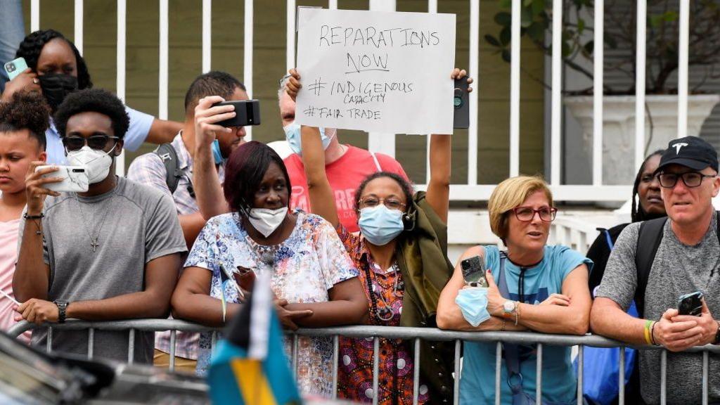 A woman holds a sign in protest as she waits for the arrival of Britain's Prince William and Catherine, Duchess of Cambridge, on the seventh day of their tour of the Caribbean on March 25, 2022 in Nassau, Bahamas