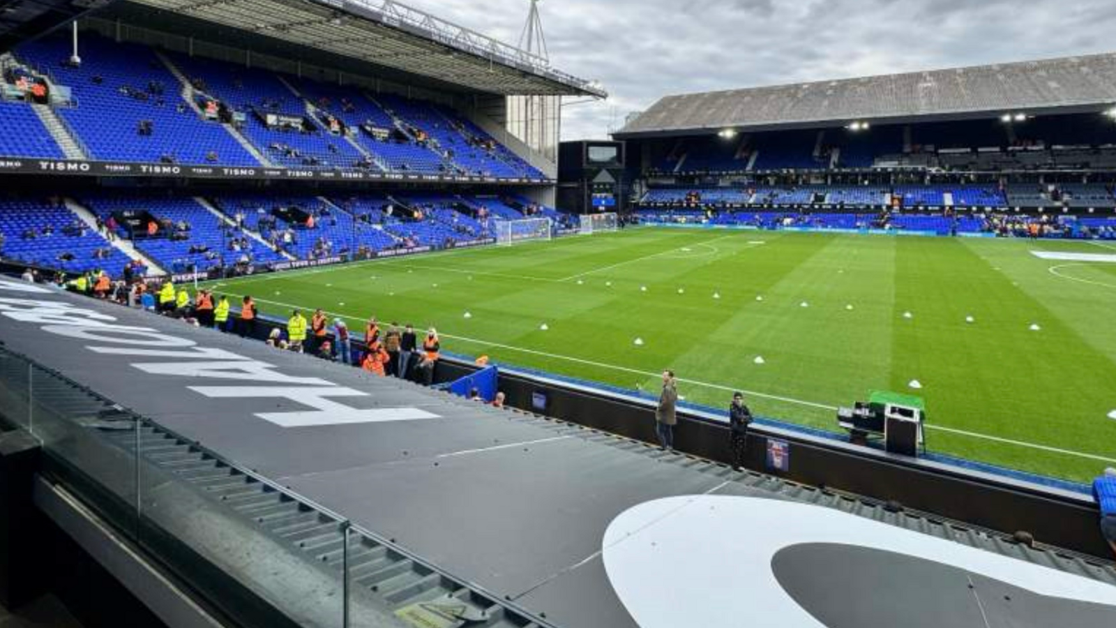 A football stadium with a few spectators in their blue seats. In the foreground is the lower row of seating of the upper tier of a stand, with a glass barrier in place between the seats and the roof of some boxes.