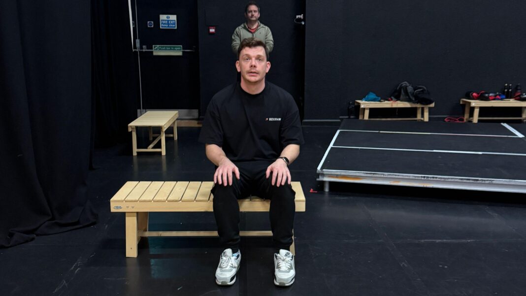 How boxing inspired a play about men’s mental health A man sitting on a bench, wearing all black looking above himself to a crowd behind the camera