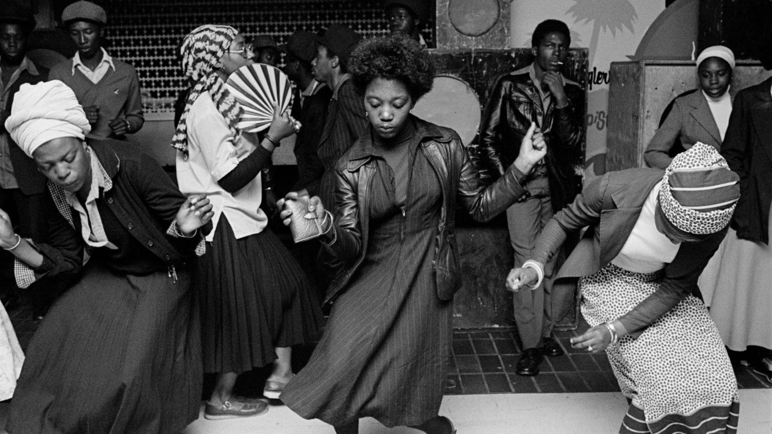 Black-and-white photograph of a crowded dance floor at Wolverhampton's Culture Centre in 1978. Several people dance to reggae music, with a central figure in a leather jacket moving across the floor while others sway and gather around the sound system behind them.