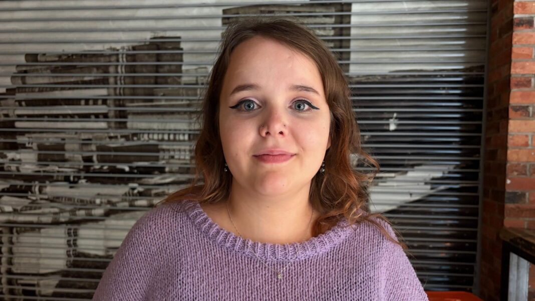 Rebecca, wearing a purple jumper and looking at the camera. She has long dark wavy hair and wears winged eyeliner.