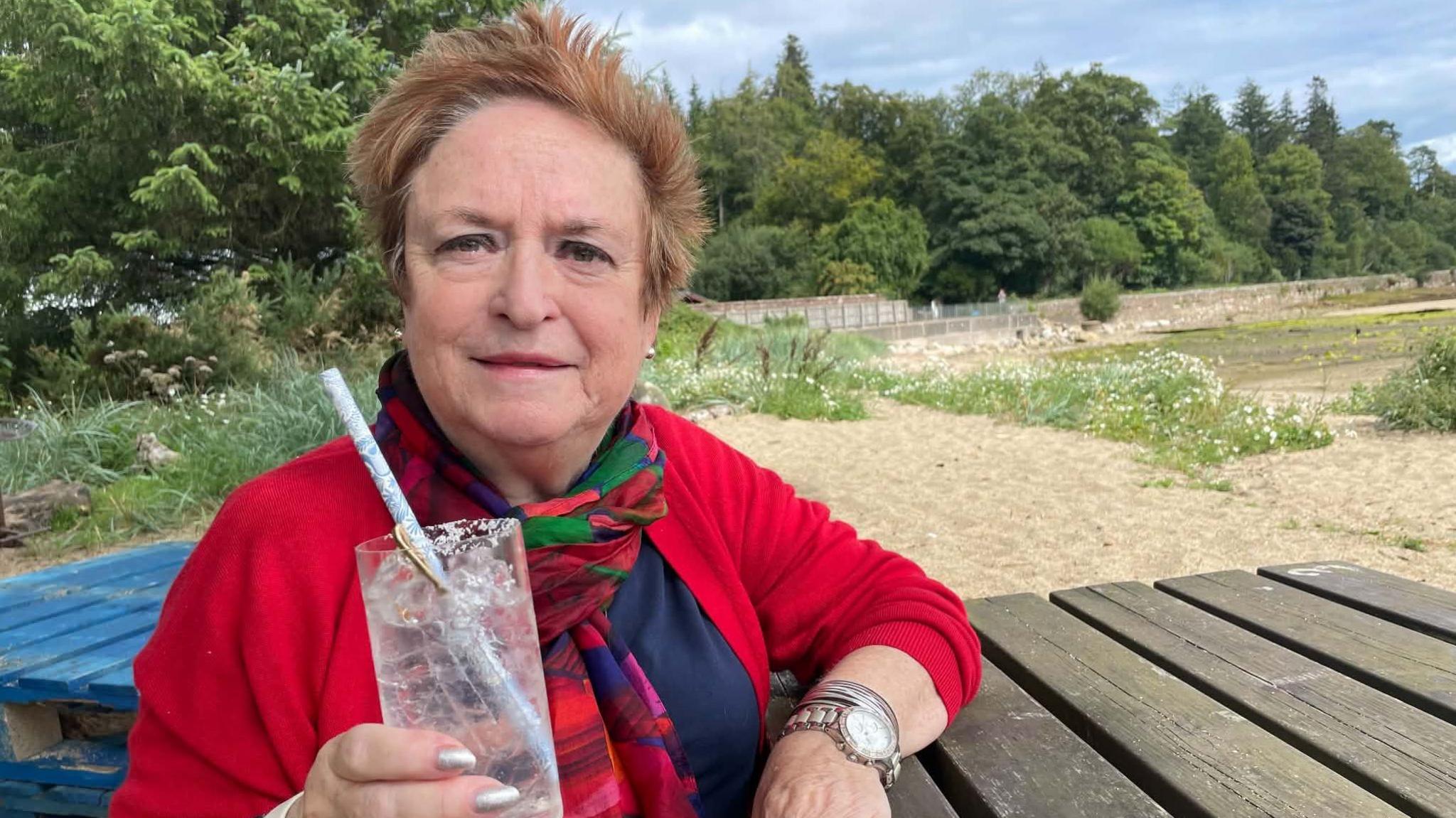 Theatre director Rosemary Hill seated at an outdoor table holding a drink beside a riverside setting. She wears a red cardigan.
