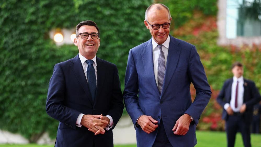 Andy Burnham smiles next to Friedrich Merz, both in navy blue suits, with green foliage in the background.