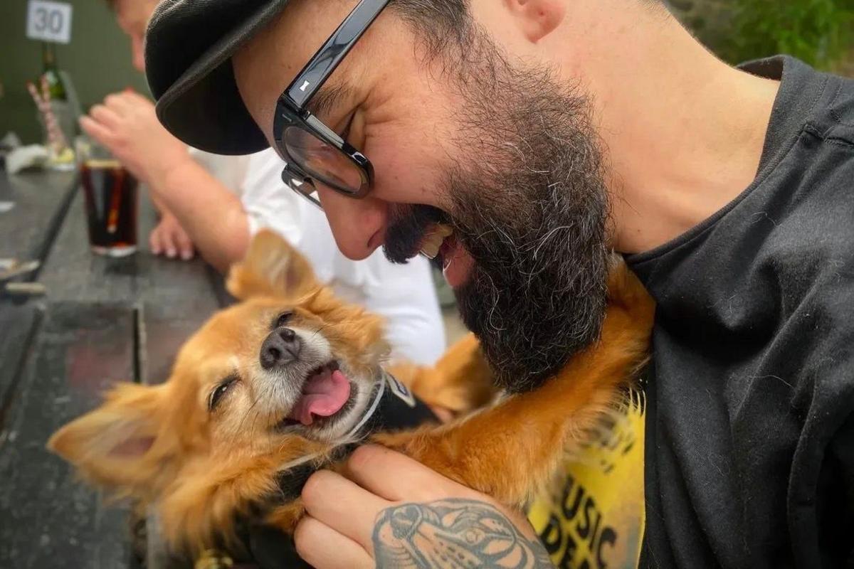 Gav Murphy has a black beard and is wearing a hat and glasses. He's holding his chihuahua-like dog up in his arms, who has her tongue out.