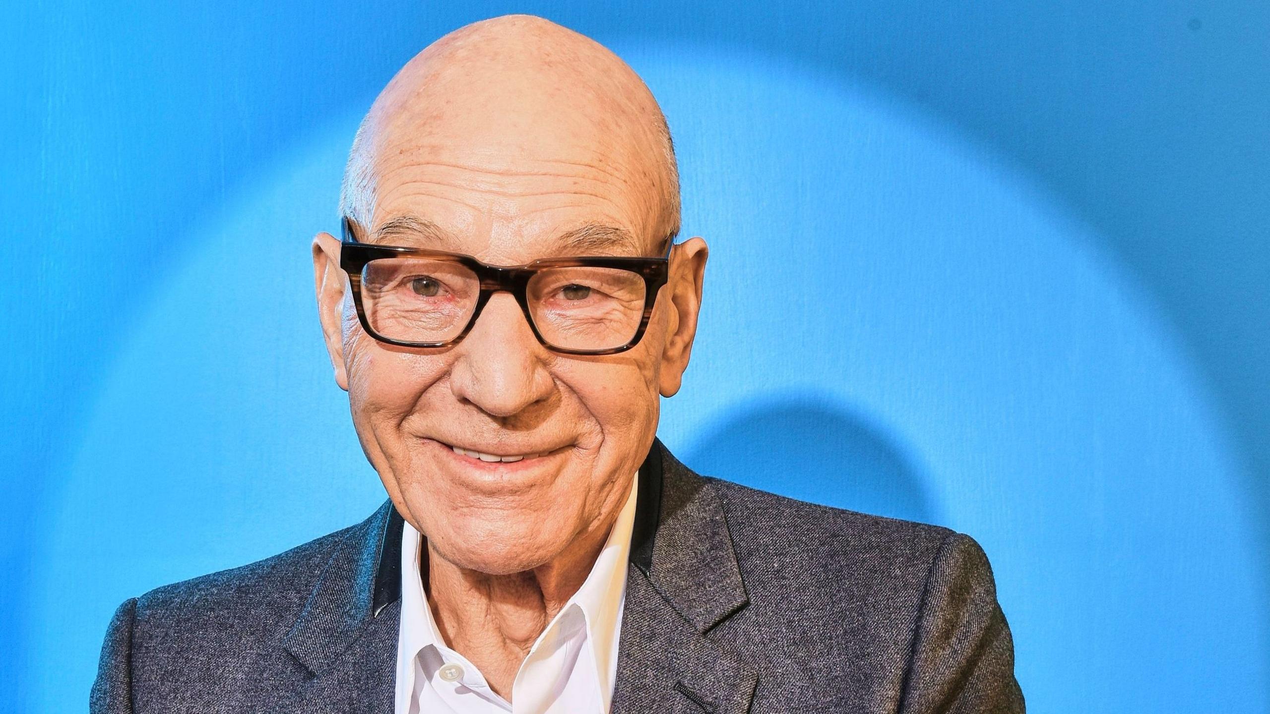 Sir Patrick Stewart smiling in a suit against a blue background in a portrait from 2022
