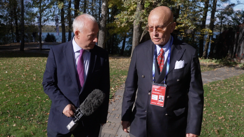 Steve Rosenberg (left) walks through a park interviewing Sergei Karaganov (right), honorary chairman of Russia's Council for Foreign and Defence Policy. Both men are in dark blue suits. Steve is holding a microphone, while Sergei has a red lanyard around his neck.