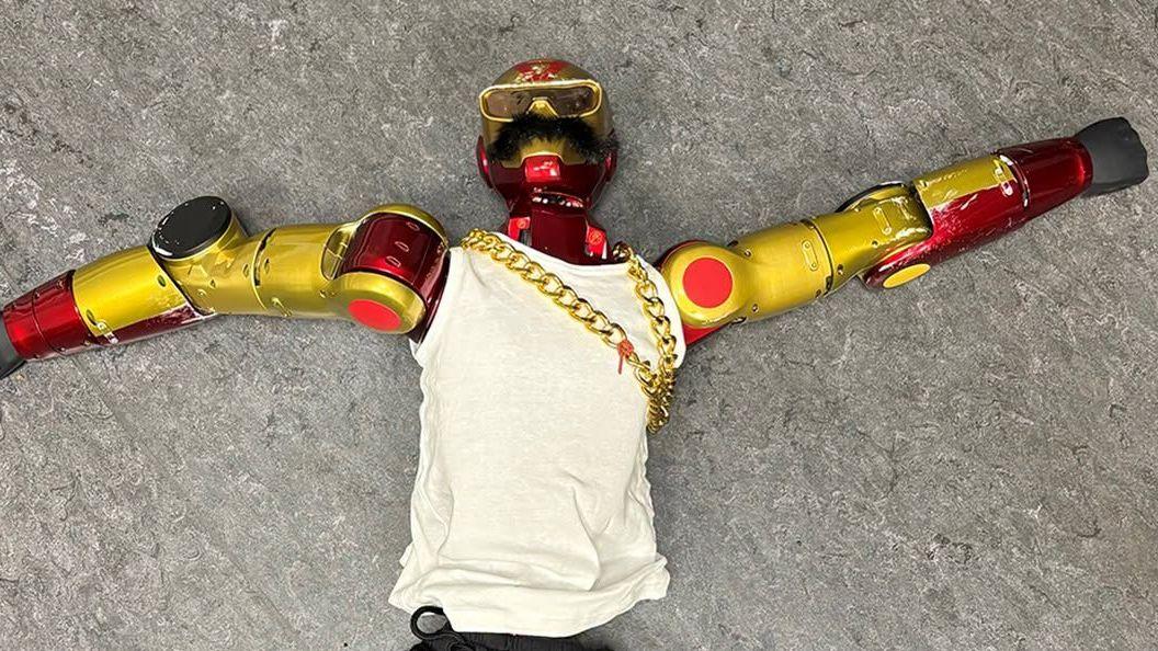 A red and gold robot with a black moustache, gold chain and white vest lying on the ground.