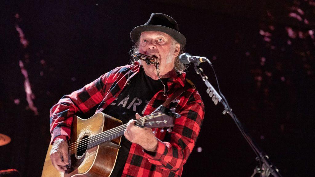 Neil young wears a red and black checked shirt and plays a guitar.