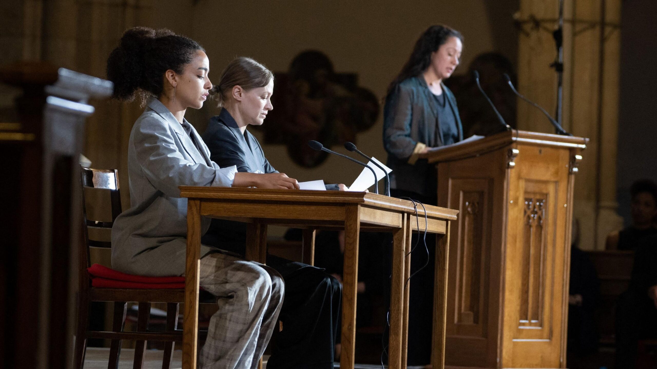 Three actresses in suits, with one standing at a lectern and two seated at a desk