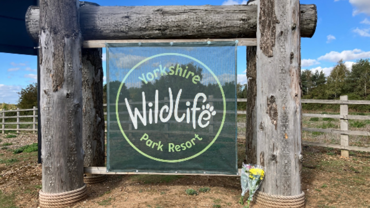The park sign with some flowers laid by it. The sign is green fabric hung on a large wooden frame. There is grass all around, There are two bunches of flowers at the scene.