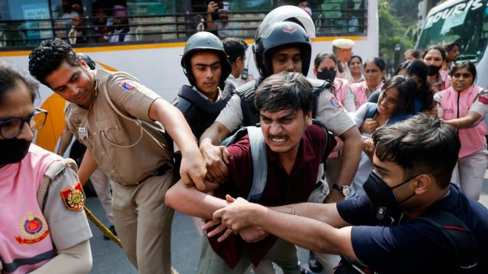 A man is being held by men in official uniforms as another man grapples with them. More people are stood behind in pink uniforms holding other protesters.