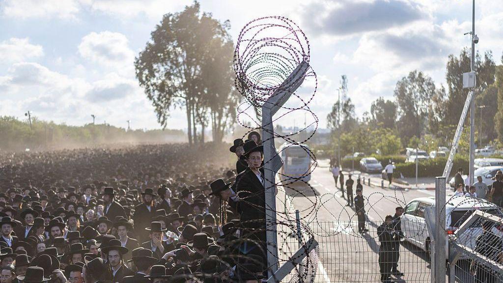 Hundreds or thousands of ultra-Orthodox jews stand beside barbed wire fencing.