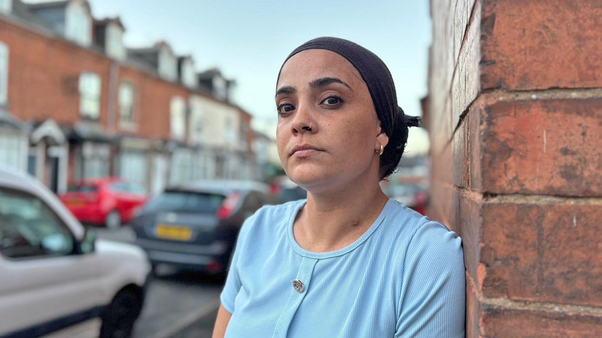 Alysha, who has a nose ring and a sky blue top, looks at the camera while leaning against a wall in Birmingham.