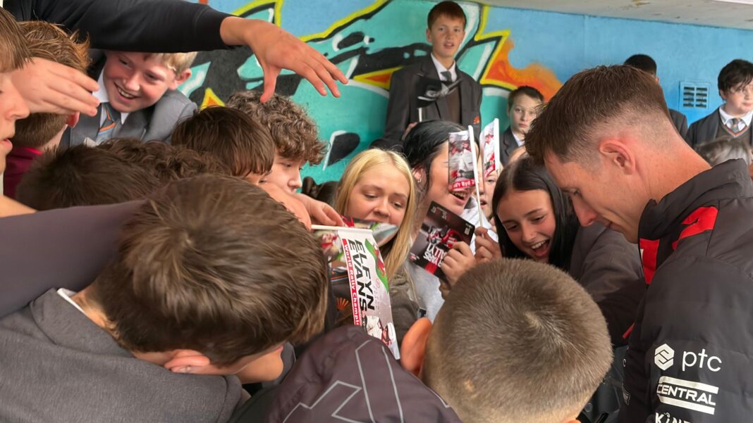 Rally champion Evans gets hero’s welcome in home county A man with brown hair wearing a black sports jacket and surrounde dby children in grey school uniforms, some holding flags with the name Elfyn Evans on it.
