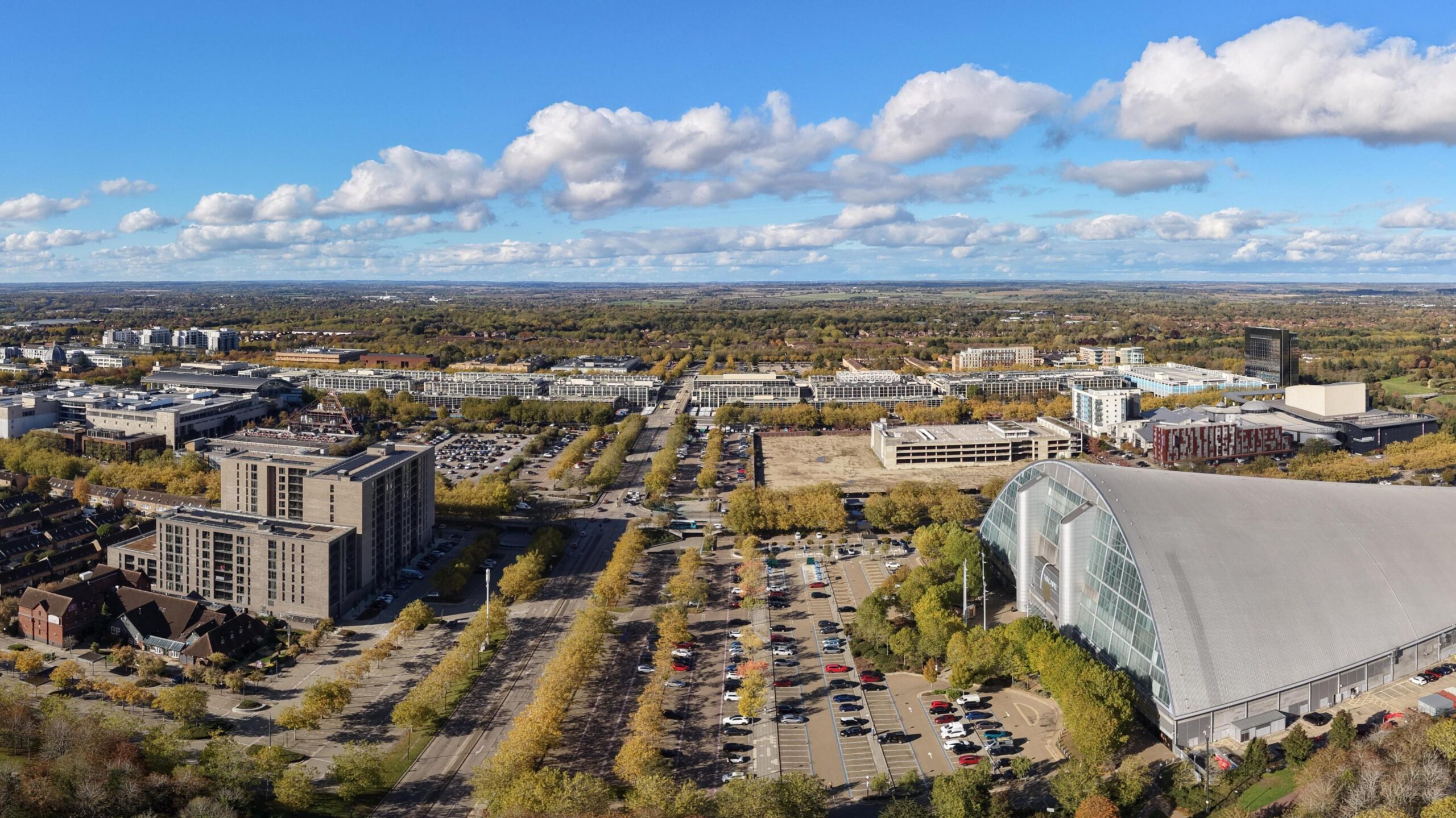 A panoramic aerial view of Milton Keynes, showing its grid roads, tree-lined boulevards and modern city centre.