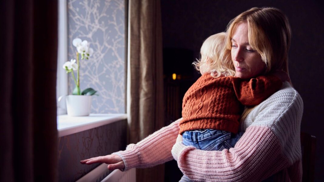 A woman in a pink and white jumper holds her child in jeans and a orange jumper while putting her hand above the radiator to feel the heat coming from it