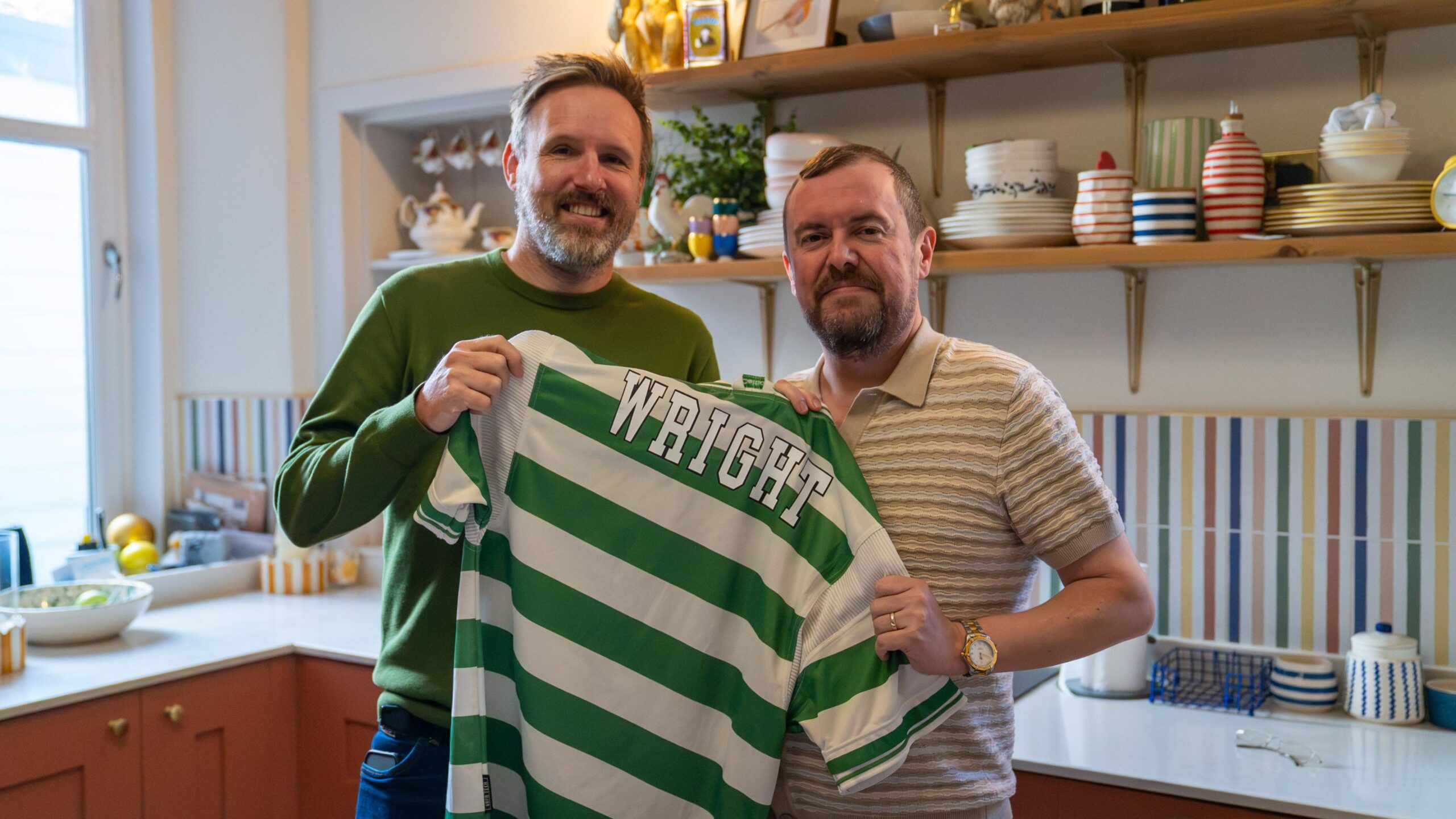 Kris and Kevin stand in Kevin's kitchen, holding up the Celtic jersey with Ian Wright's name on it.