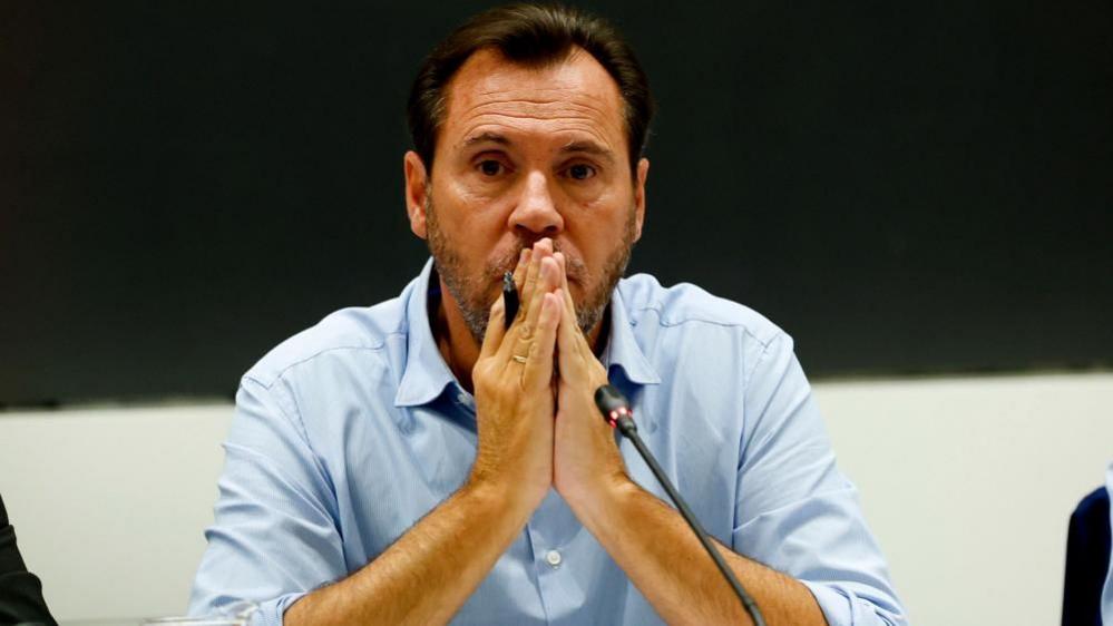 A middle-aged man in a light blue button-down shirt, placing his hands together in a prayer position in front of his mouth while sitting in front of a microphone at a press conference.