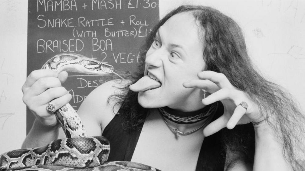 Conrad Cronos Lant in a black and white publicity shot from 1985. He is holding a large snake in his right hand so the snake is looking at him with its tongue out and Lant is looking back at the snake with his tongue out. Lant has thinning but long and wispy dark hair. His left hand is raised in a claw shape and he has a waistcoat on over bare skin. The snake is curled up on a table and behind them is a chalkboard with items written on it as if it is a menu board in a cafe. These include "Mamba + Mash £1.30" and "Snake Rattle + Roll (with butter)".