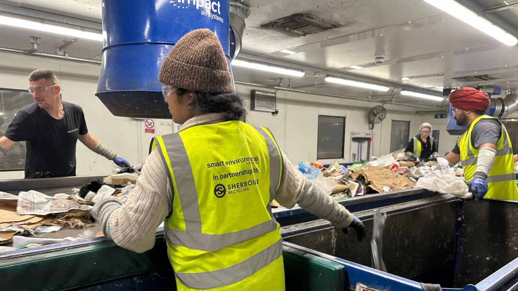 Four workers wearing high-visibility safety vests, gloves and protective eyewear sort mixed waste and recyclables on a conveyor belt inside a recycling facility. Large blue extraction equipment hangs above the sorting line, while a strip lights illuminate the industrial workspace.