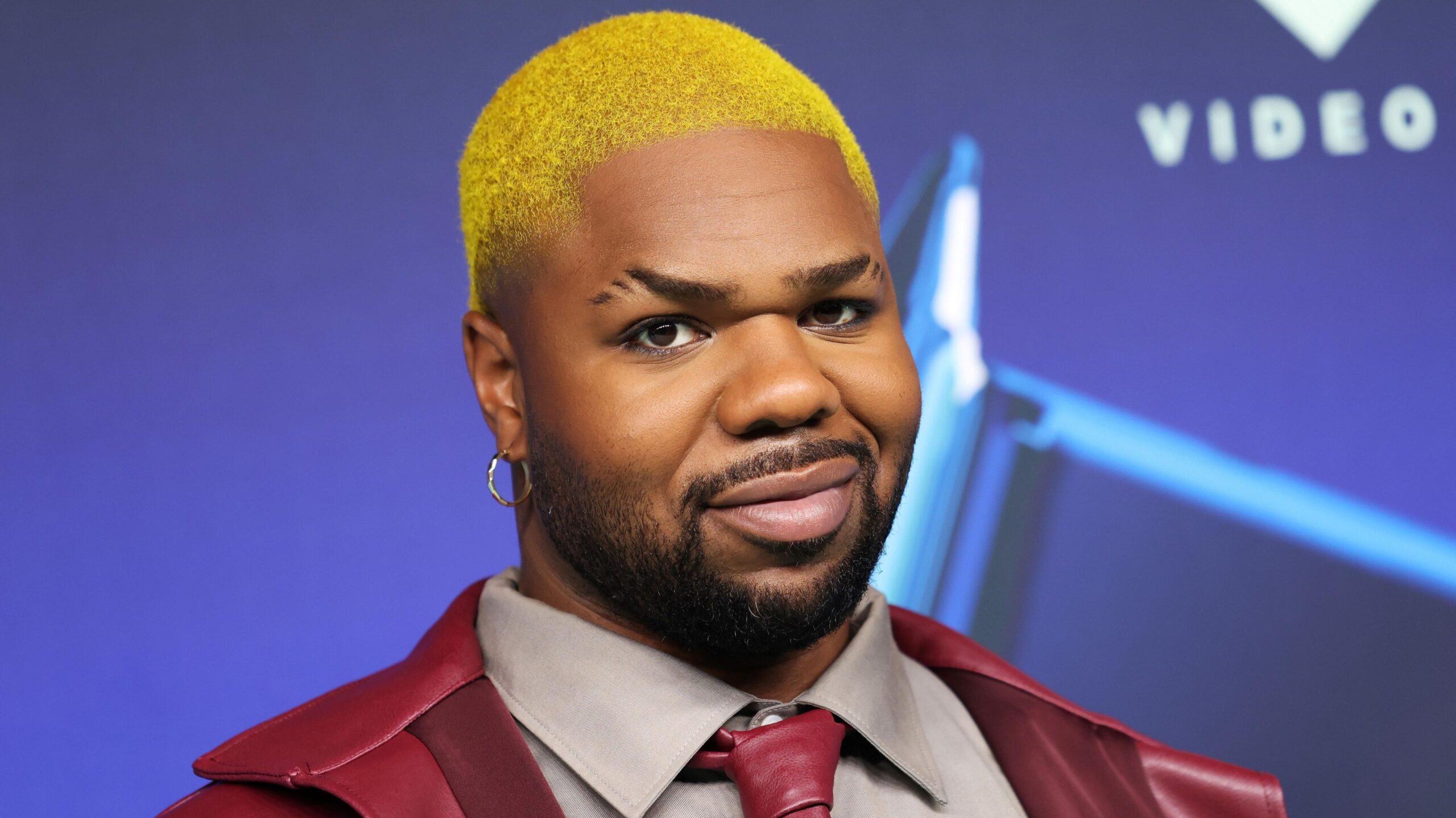 MNEK wears a red leather jacket over a grey shirt with a red leather tie. He has short cropped yellow hair.