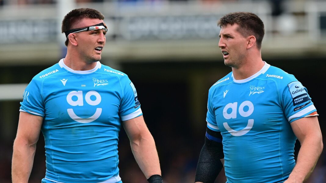 Tom Curry (left) stands looking to his left towards brother Ben Curry. Both are wearing light blue Sale Sharks kit from 2024