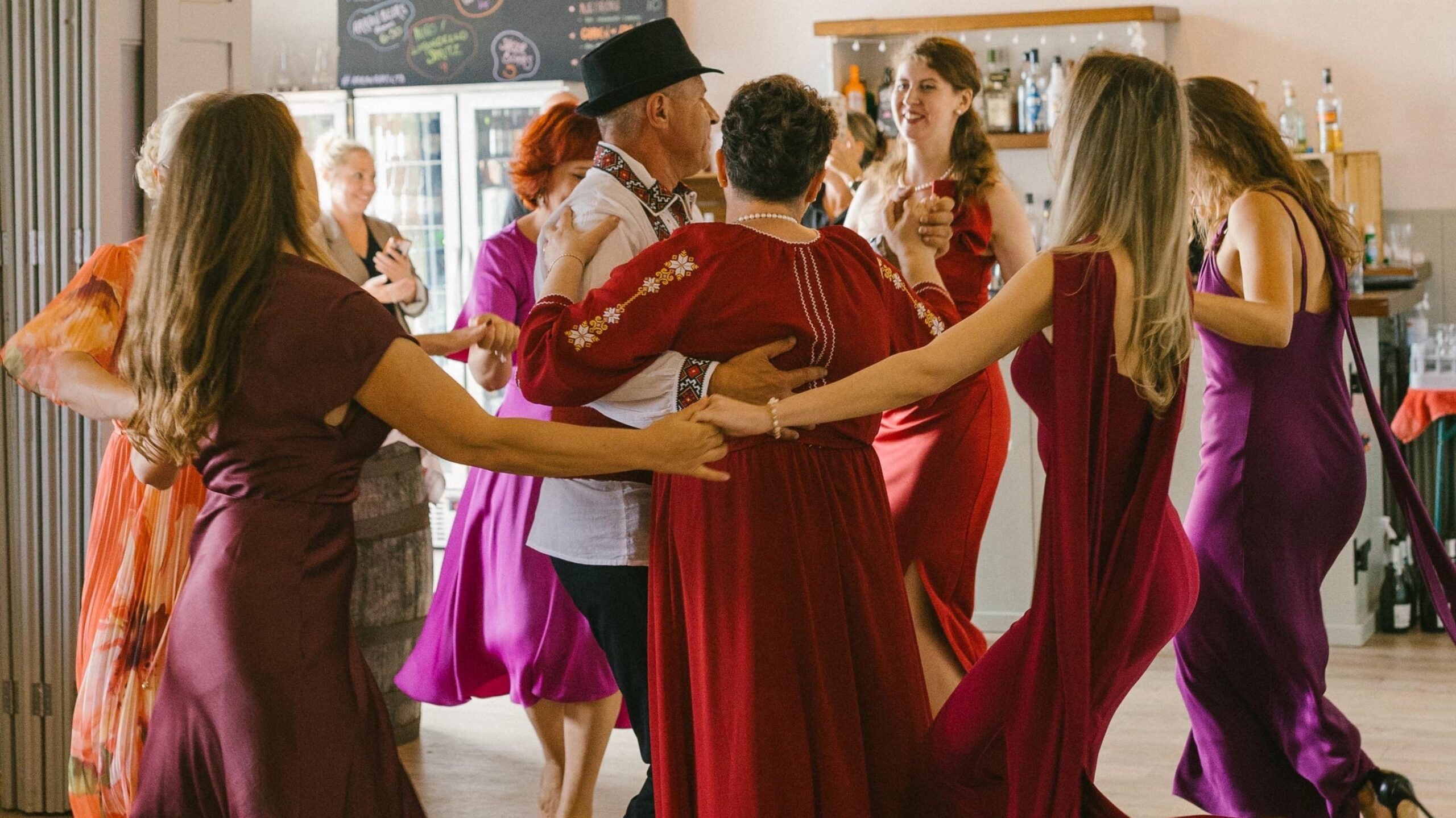 An older couple dressed in traditional Moldovan attire stand in the centre of a ceilidh circle. The woman wears a long red dress that has long sleeves with embroidery down the arms. The man wears a black short brimmed hat and a white shirt that is embroidered down the collar and round the cuffs of the sleeves. The woman dancing in a circle around them wear dresses of red and purples.