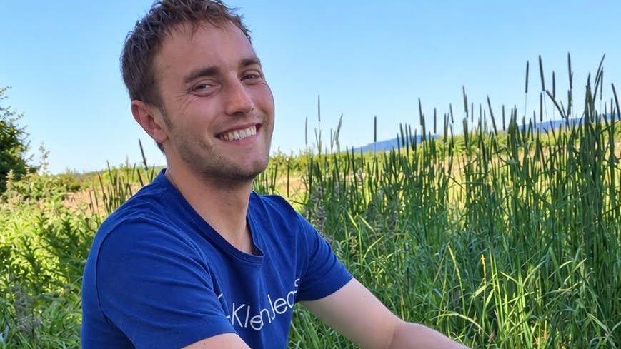 Hugo Flint Cahan is looking into the camera smiling. He is sat in a field surrounded by long grass on a bright summer day. He is wearing a blue t-shirt.