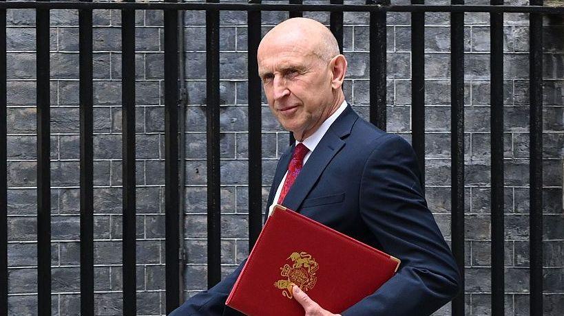 Chancellor John Healey walks outside, wearing a navy suit with a red tie. He is holding a red folding and is looking to the side. Behind him is a brick wall and black railings.
