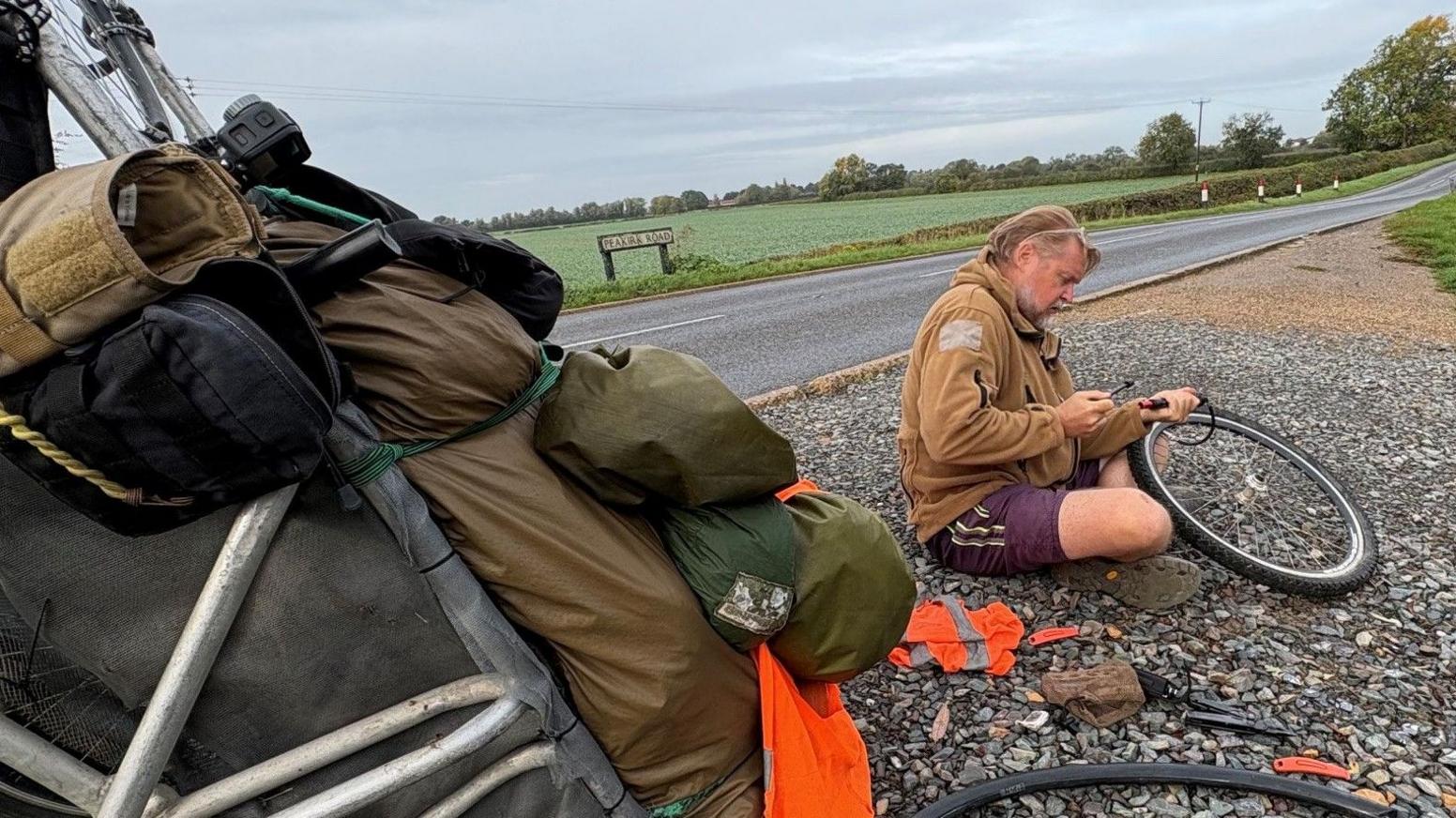 A man in a mustard coloured fleece and shirt sits by the roadside repairing a punctured tyre on his handcart. The card is laden with essential equipment.