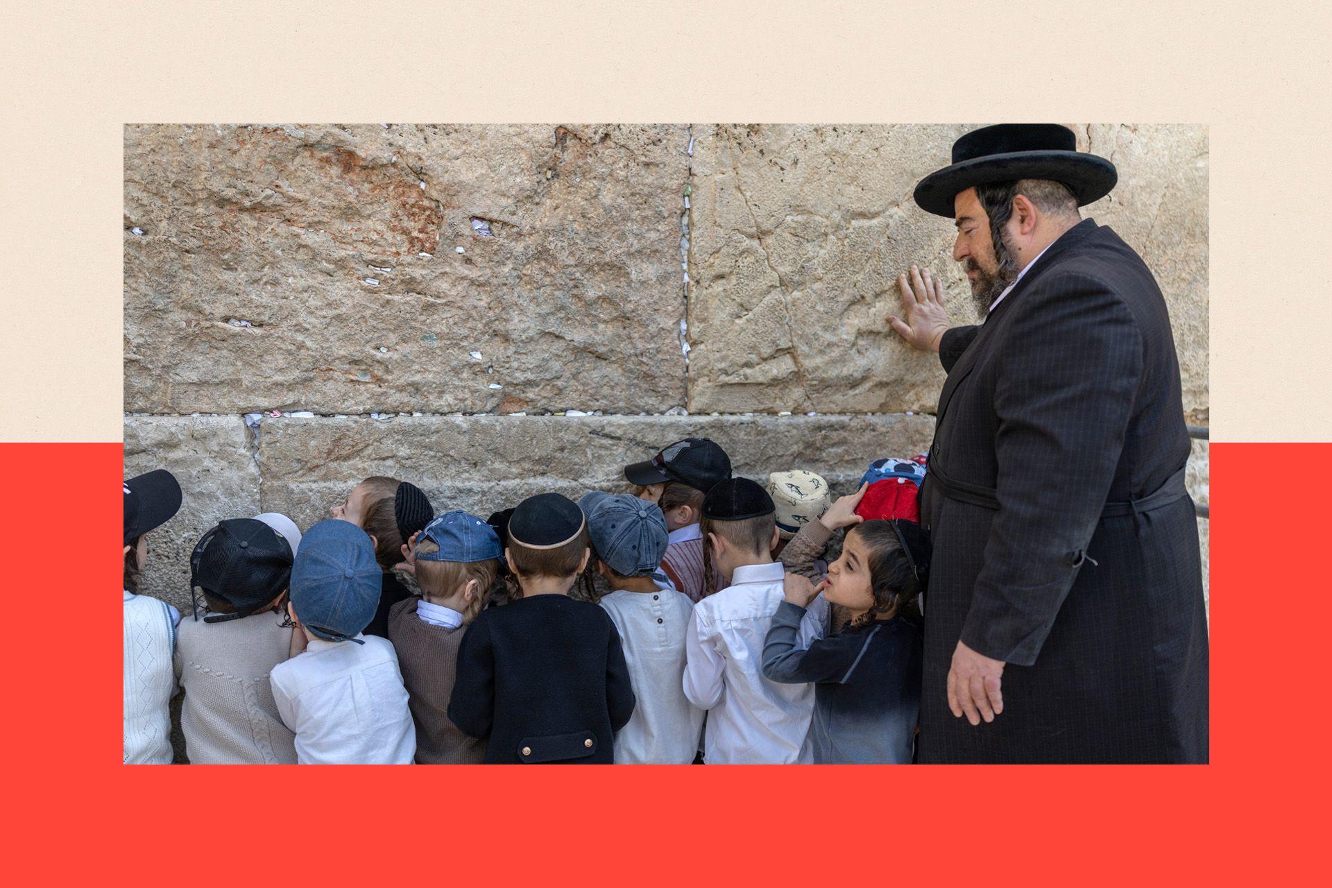 A group of ultra-Orthodox boys pray at the Western wall, with their backs to the camera. A Haredi man stands with them