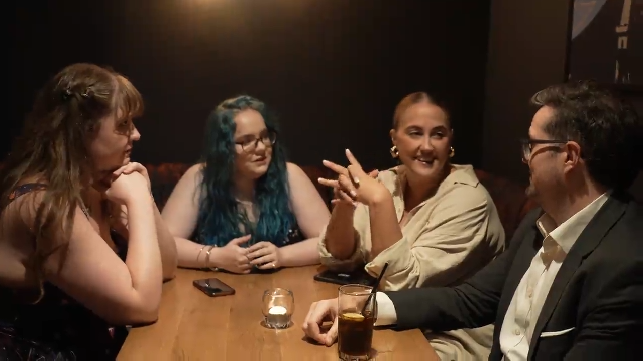 Olivia sits at a table in a bar with two other women and a man. He has a drink in front of him and there is a tealight in a small glass in the middle of the table