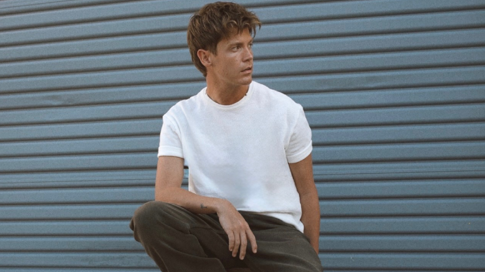 A young man with light brown hair. He is wearing a white t-shirt and dark trousers and is looking off to the side, with one arm resting on one knee.