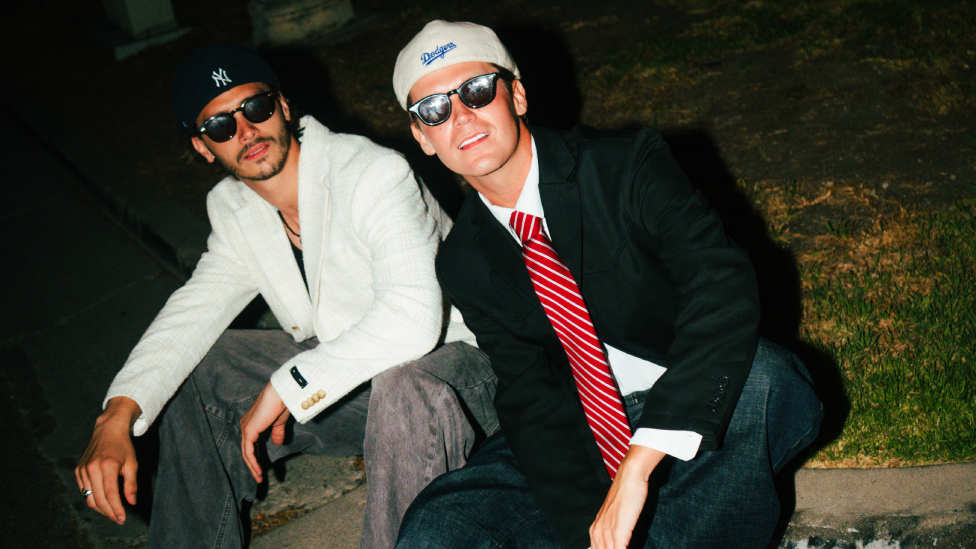 Two young men, sat next to each other on a pavement, both wearing baseball caps, sunglasses, jeans and blazers and looking at the camera. The one on the left has dark hair and facial hair and a white blazer and black t-shirt. The one on the right has a black blazer, white shirt and red striped tie and is clean shaven with short hair.
